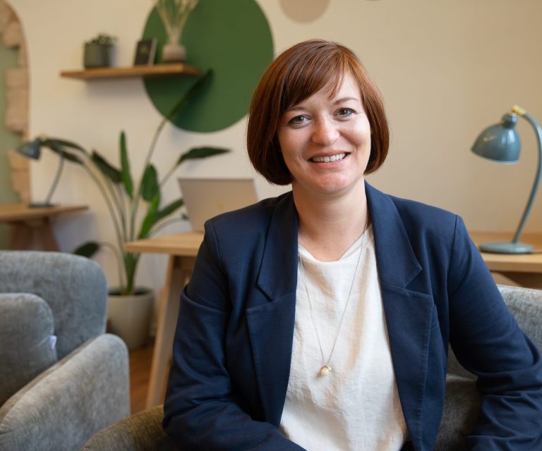 Birgit in einem blauen Blazer, lächelt in einem modernen Büro.