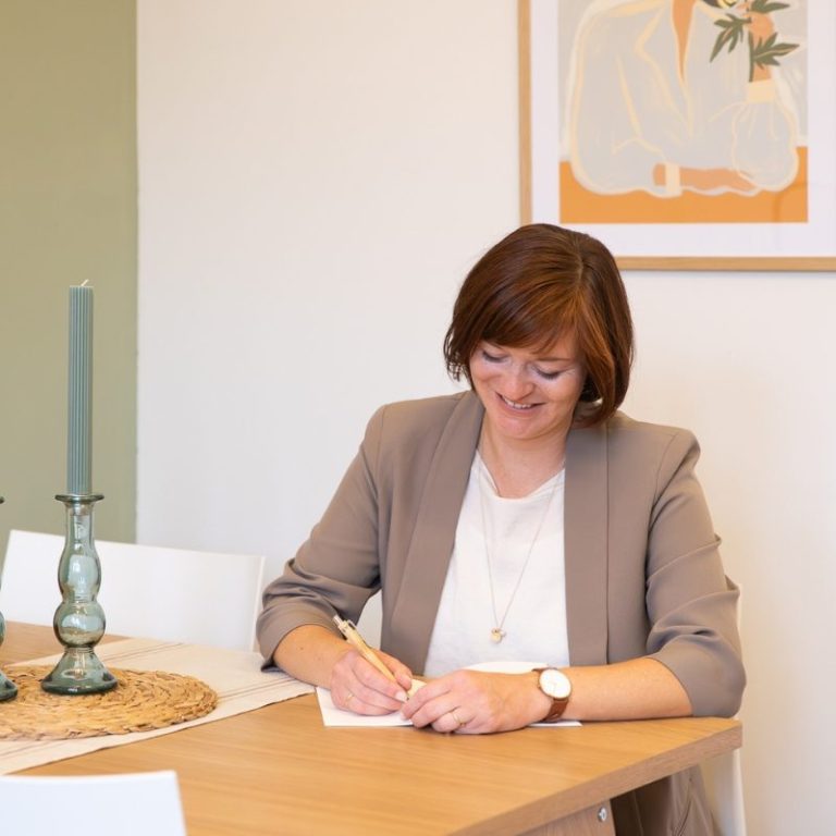 Birgit Gebauer sitzt am Tisch, schreibt in ein Notizbuch, dekorativ mit Kerzen und Kunstwerk im Hintergrund.