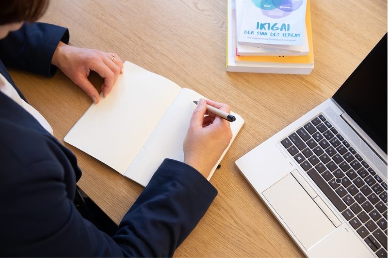 Porträtfoto Birgit Gebauer (Eva Korn) Birgit Gebauer schreibt in ein Notizbuch neben einem Laptop, berufliche Orientierung, Ikigai, Vision