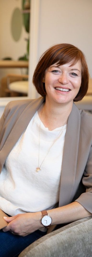 Birgit Gebauer in beigem Blazer, sitzt entspannt in einem modernen Raum, offen, einladend.