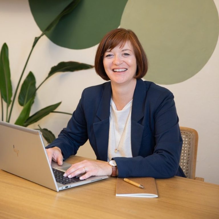 Birgit am Computer lächelt am Schreibtisch, grüner Pflanze im Hintergrund.