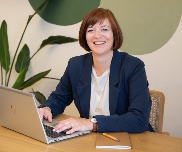 Porträtfoto Birgit Gebauer (Eva Korn) Birgit Gebauer sitzt an einem Tisch mit Laptop, lächelt und hat Notizbuch neben sich, Online-Coaching, AVGS-Coaching