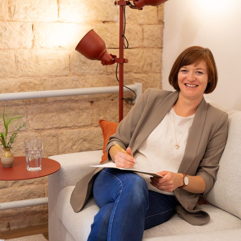 Birgit Gebauer sitzt auf einem Sofa, lächelt und hält einen Stift in der Hand, mit Tisch und Lampe im Hintergrund.